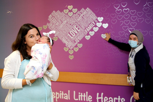New mum holding her baby in front of the hospital’s baby wall, along with a staff member holding a love heart against the wall
