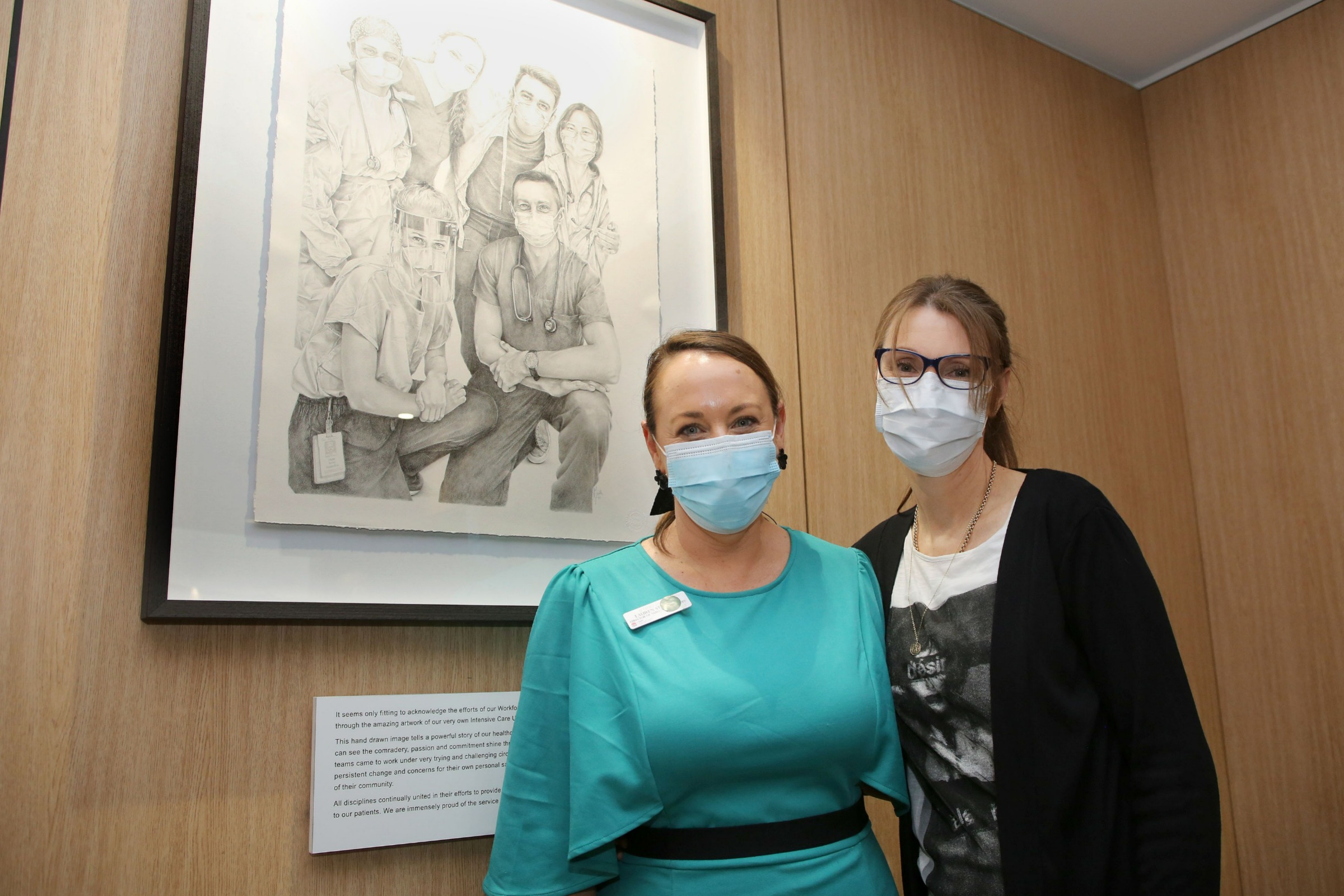 Lauren Sturgess and Jennifer North stand in front of a pencil drawing of hospital staff during the COVID-19 pandemic.
