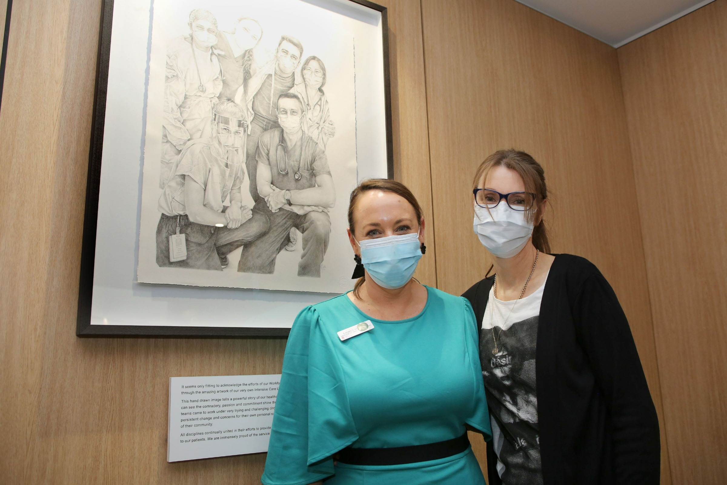 Lauren Sturgess and Jennifer North stand in front of a pencil drawing of hospital staff during the COVID-19 pandemic.