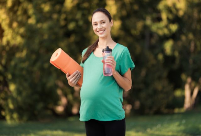 A pregnant woman smiling in a park with a yoga mat and water bottle