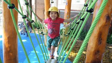child wearing yellow hat walking across a play equipment bridge