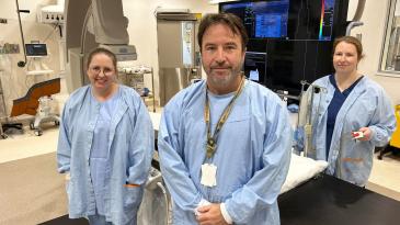 three staff members stand in the cardiac cath lab next to the CT machine